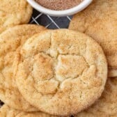 A close-up of several snickerdoodle cookies on a cooling rack, with a small bowl of cinnamon sugar in the background. The cookies are golden brown and sprinkled with cinnamon sugar.