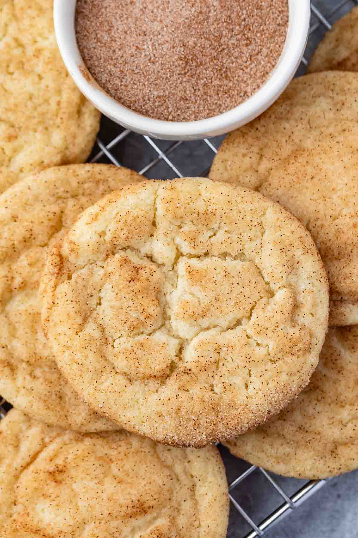A close-up of several snickerdoodle cookies on a cooling rack, with a small bowl of cinnamon sugar in the background. The cookies are golden brown and sprinkled with cinnamon sugar.
