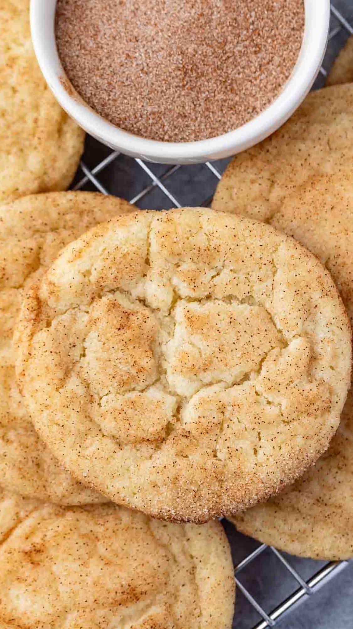 A close-up of several golden-brown snickerdoodle cookies on a cooling rack, with a small white bowl of cinnamon sugar in the background.