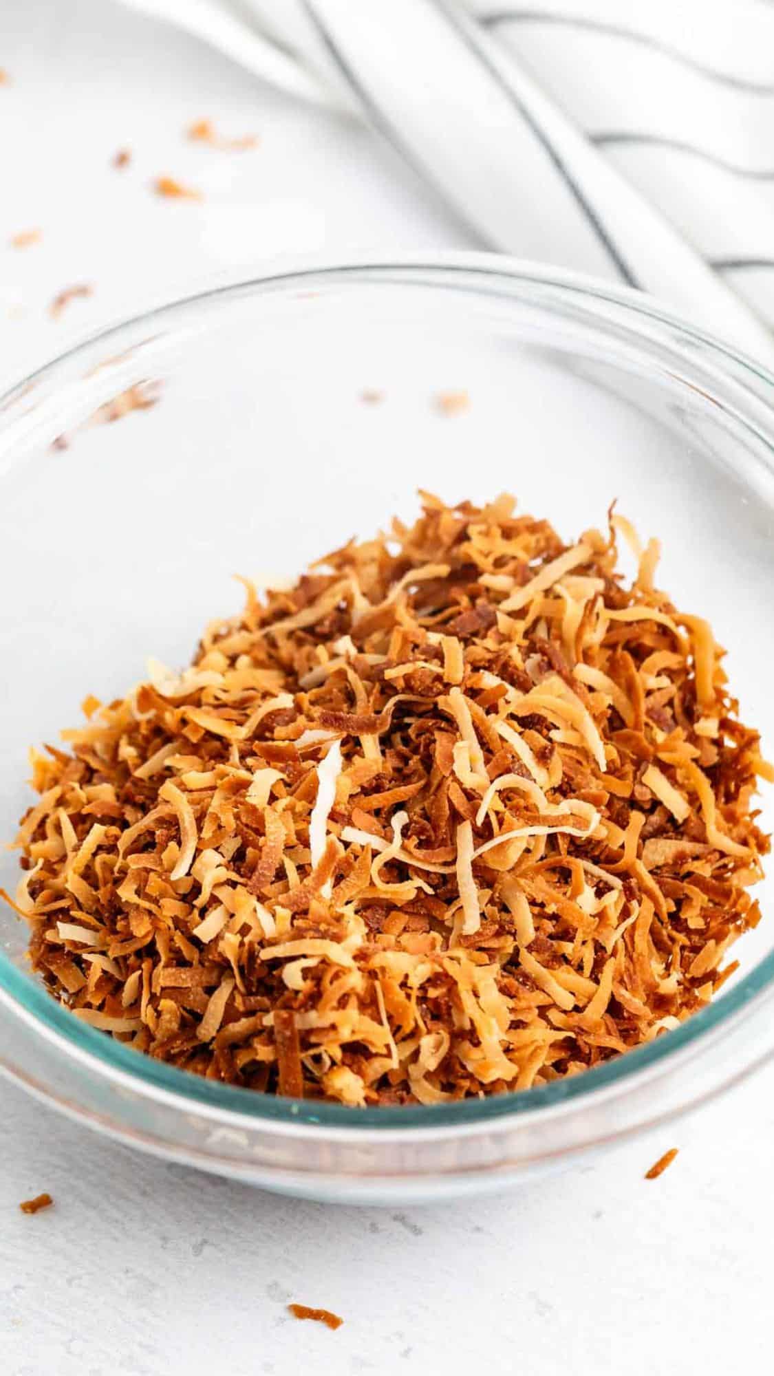 A clear glass bowl filled with golden-brown toasted shredded coconut sits on a white surface, with a striped cloth in the background and a few coconut flakes scattered nearby.